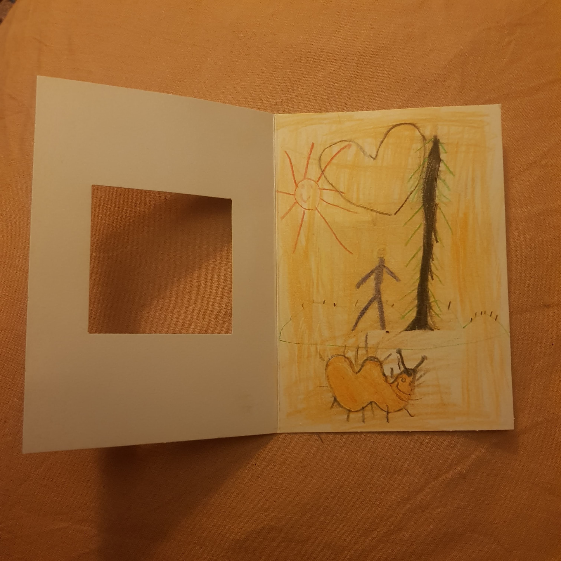Inside the card: a crayon drawing of a person standing by a tall pine tree under a sun and a heart, with a smiling orange caterpillar in the foreground