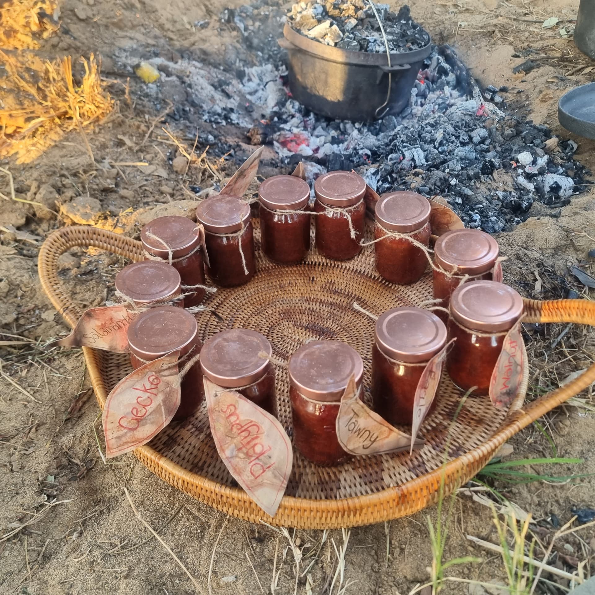 Hand-labelled jars of jam in a woven tray beside the camp fire