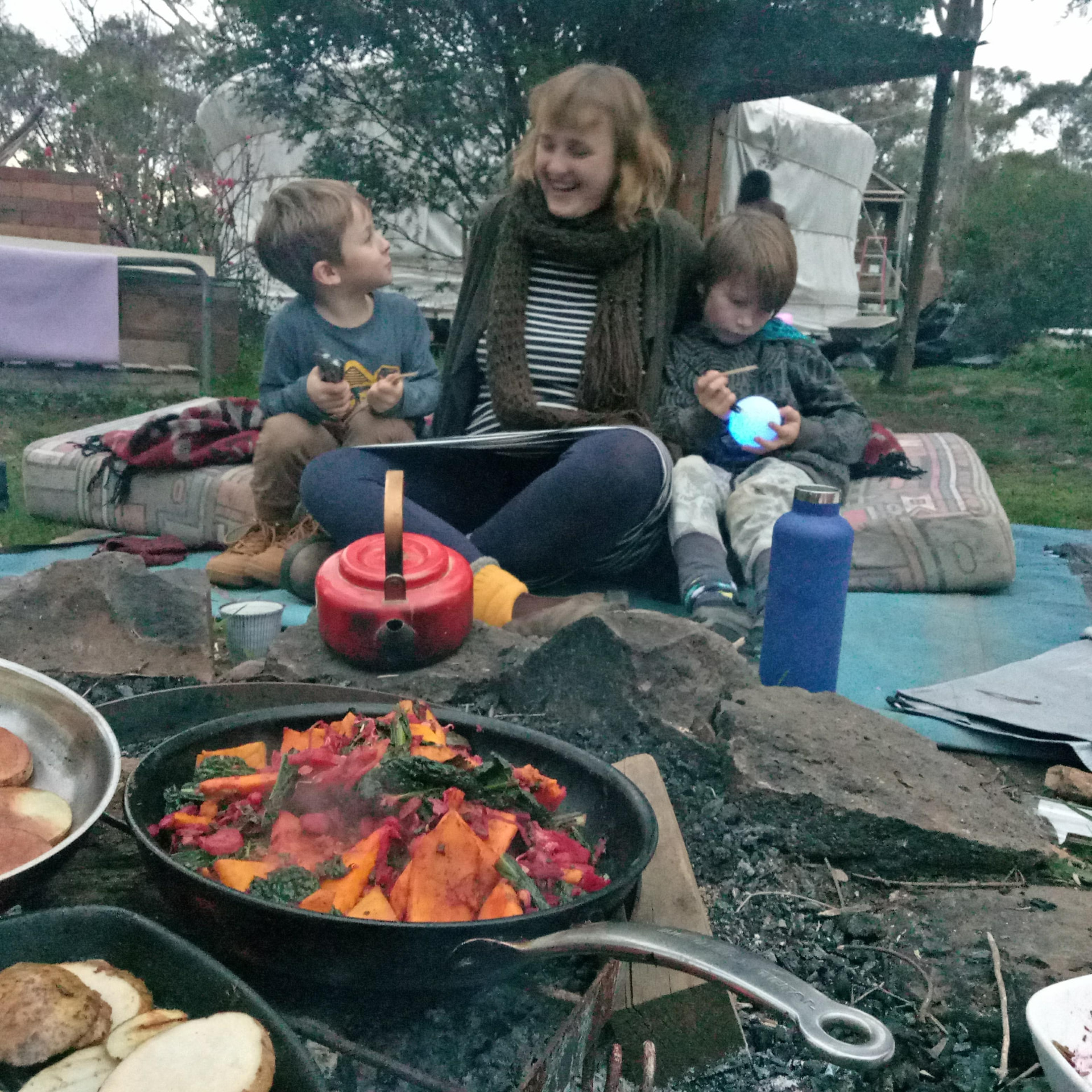 A mentor and two children beside a fire with a pan of roasted vegetables